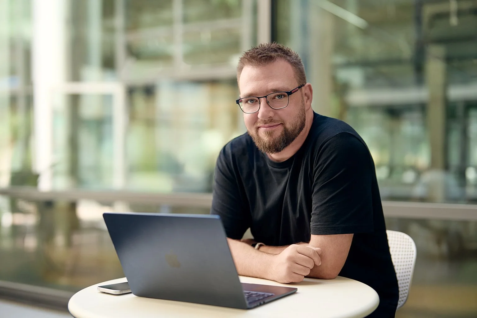 Paul Böstro sitzt lächelnd mit seinem Laptop an einem Tisch in einem hellen Raum