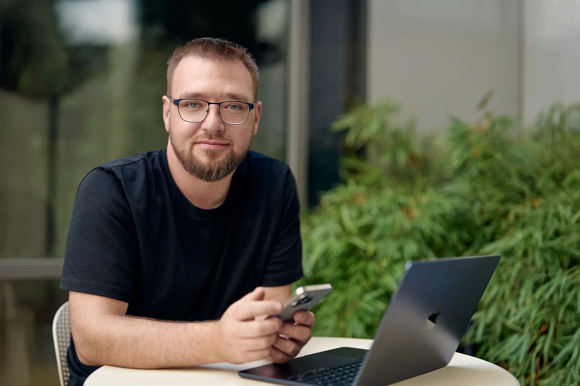 Paul Böstro sitzt draußen an einem Tisch mit Laptop und Handy, im Hintergrund Pflanzen