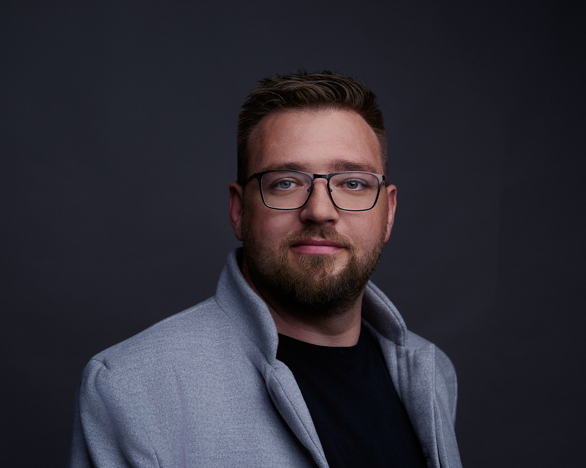 Ein Mann mit kurzem Haar, Bart und Brille trägt graue Jacke, schwarzes Shirt vor dunklem Hintergrund.
