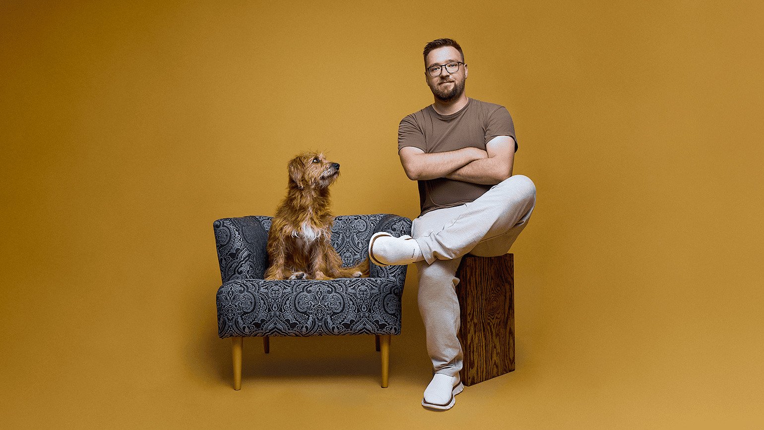 Ein Mann sitzt mit verschränkten Armen auf einem Holzblock neben einem braunen Hund auf einem gemusterten Stuhl vor gelbem Hintergrund.